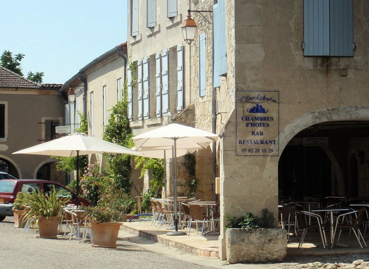 Stone buildings with pal blue shutters, an outdoor café with small potted trees and white shade umbrellas