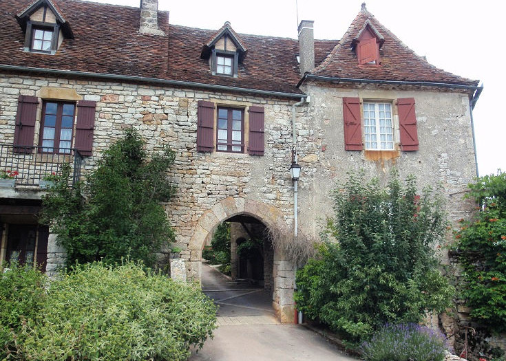 Turreted stone building which serves as a gateway to the village of Loubressac through an arched entrance. Brown shutters adorn windows on the upper level