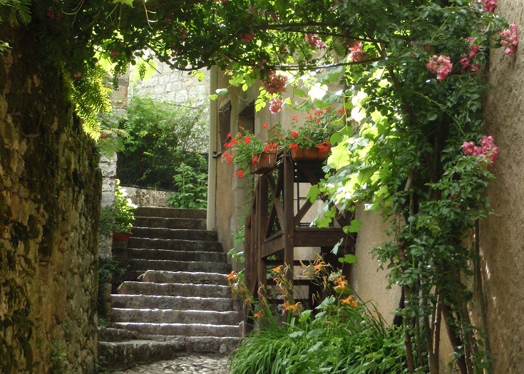 Narrow laneway with stone steps and a wooden water wheel hung with bougainvillea and other flowering plants